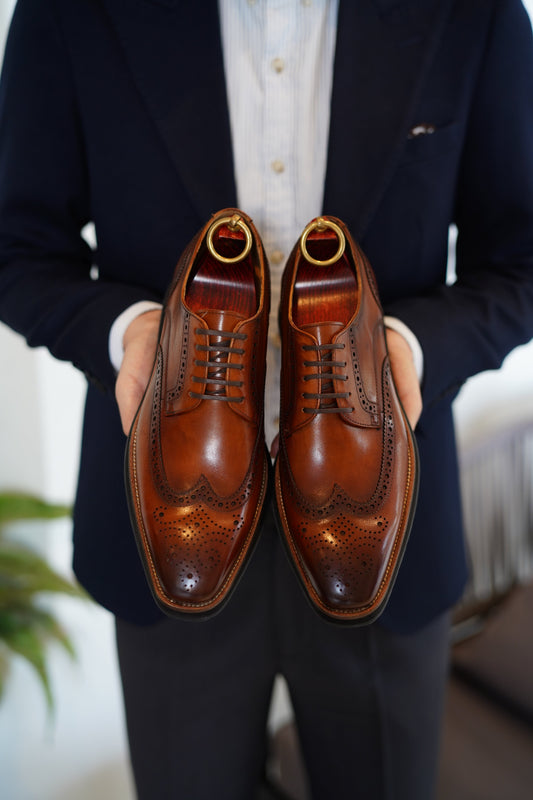 Close-up of a cognac leather brogue shoe with intricate wingtip detailing, held by a man in a navy suit.