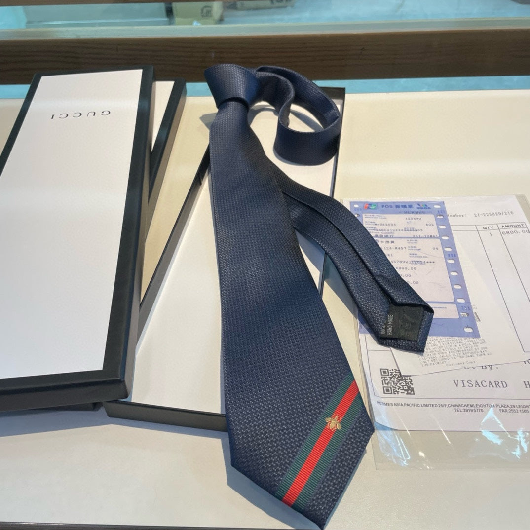 A navy blue silk tie with a green and red stripe and an embroidered bee detail, displayed on a table with a white background, next to its packaging.