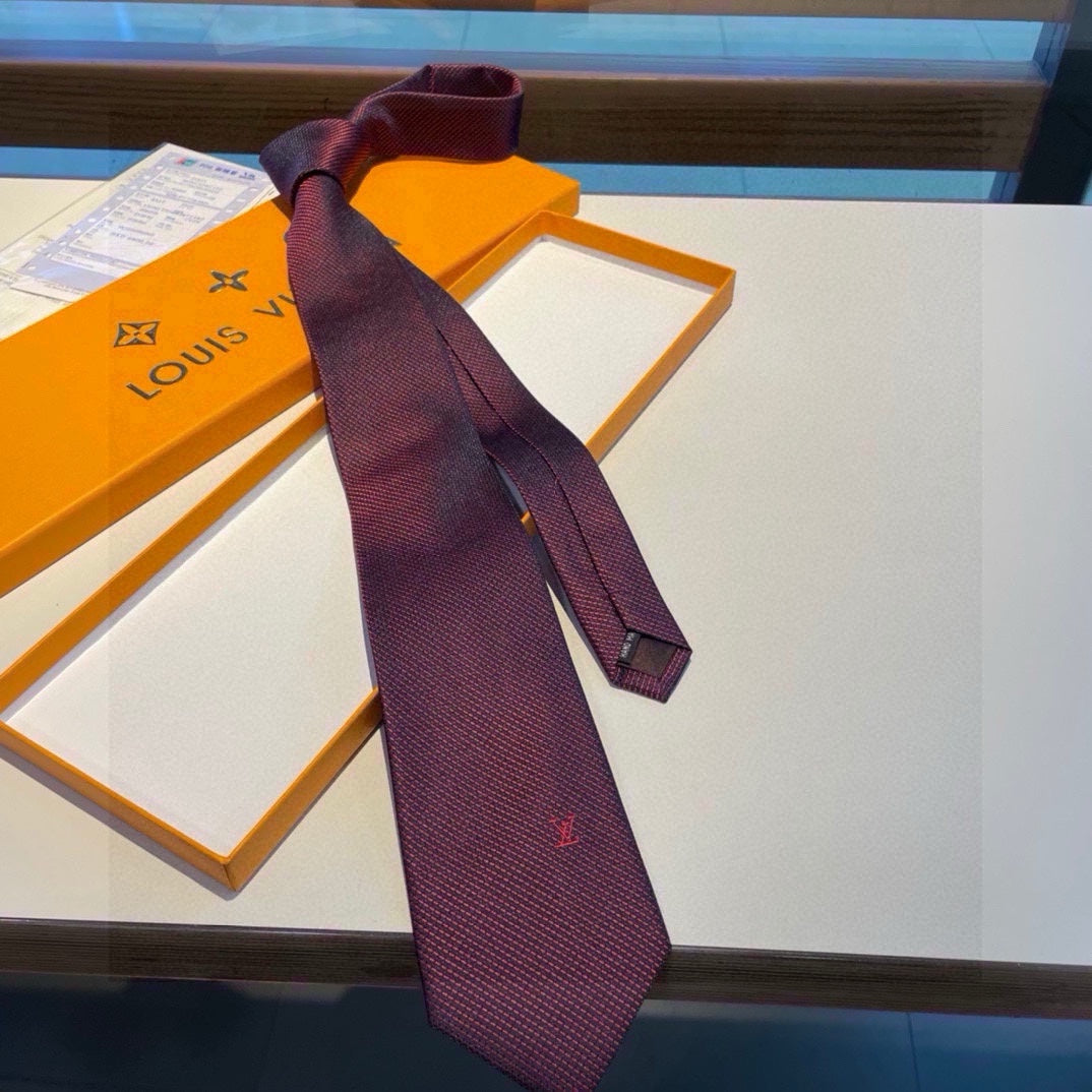 A burgundy Louis Vuitton silk tie with a subtle red micro-pattern and an embroidered LV monogram, displayed on a table next to an orange box.