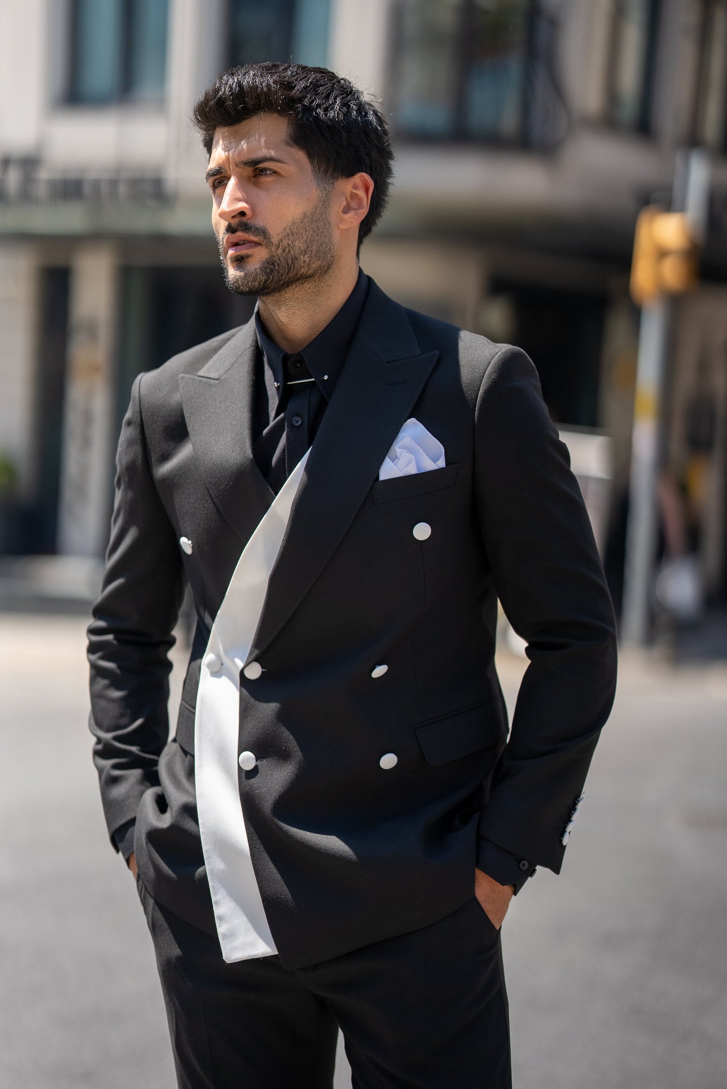 Man in a black suit with a white pocket square standing on a city street.