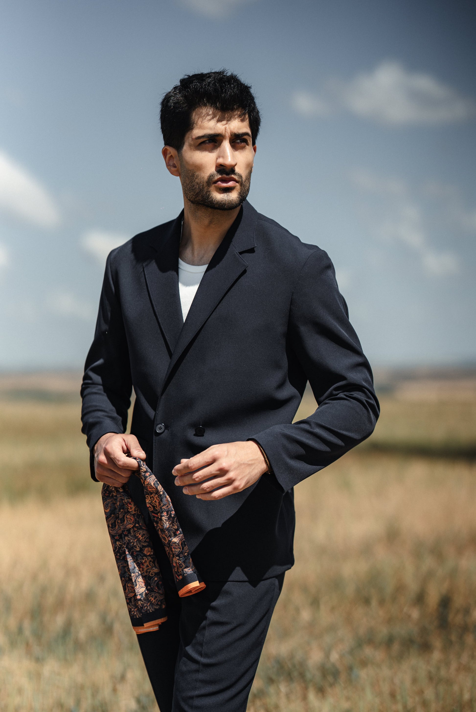 Man in a dark suit holding a decorative bottle in an open field with a blue sky.