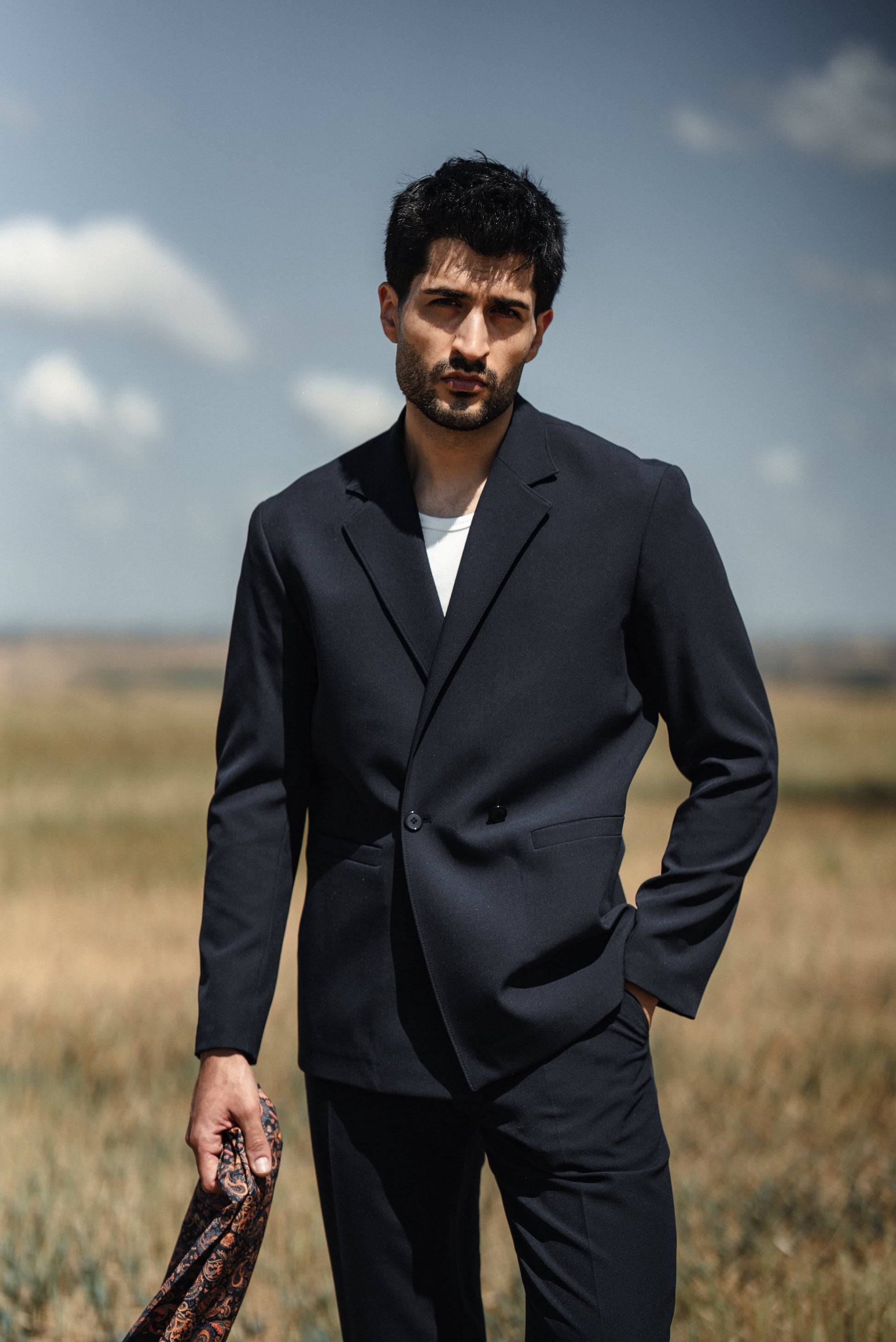 Man in a dark suit standing in an open field with a blue sky.
