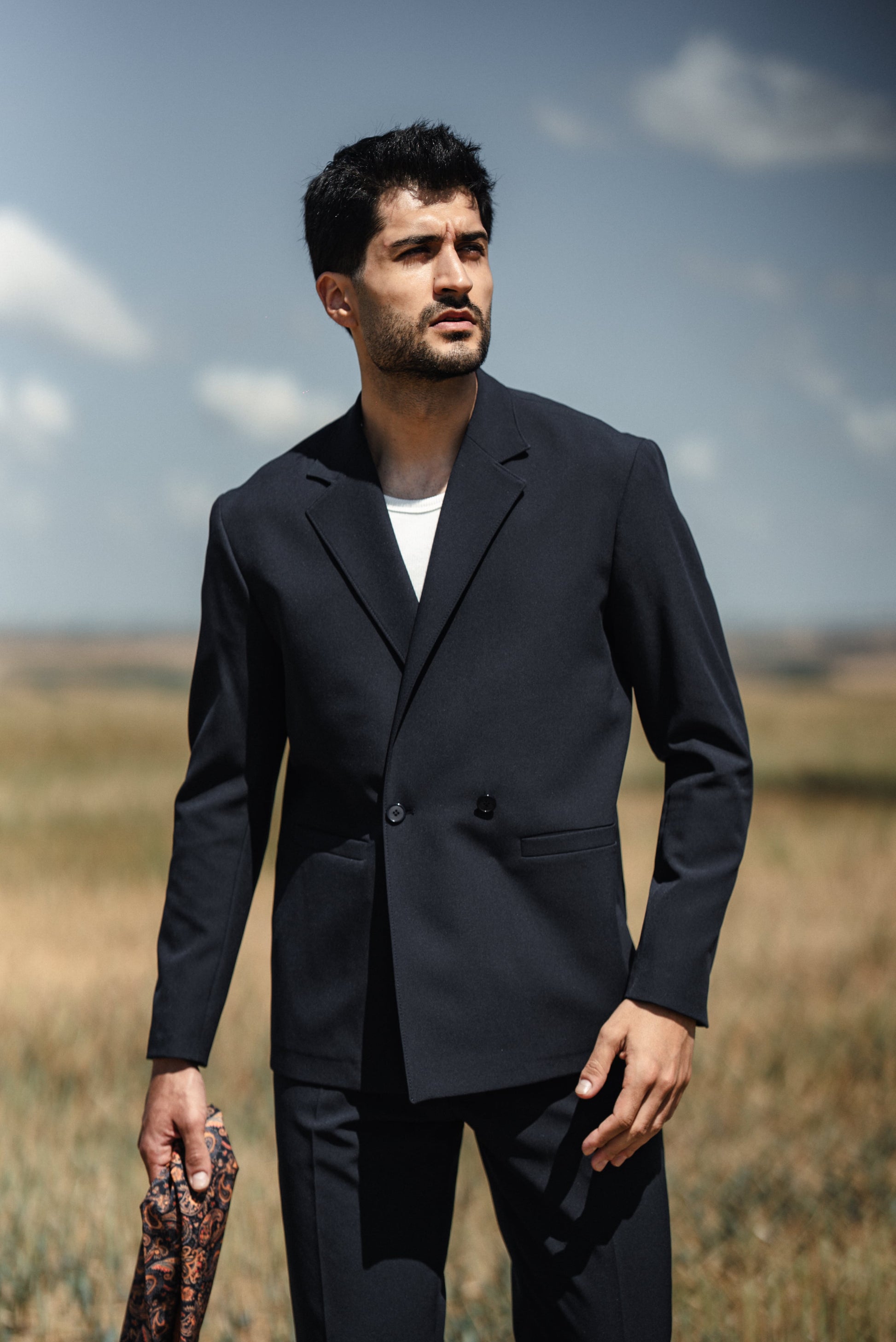 Man wearing a dark suit standing in an open field with a clear sky.