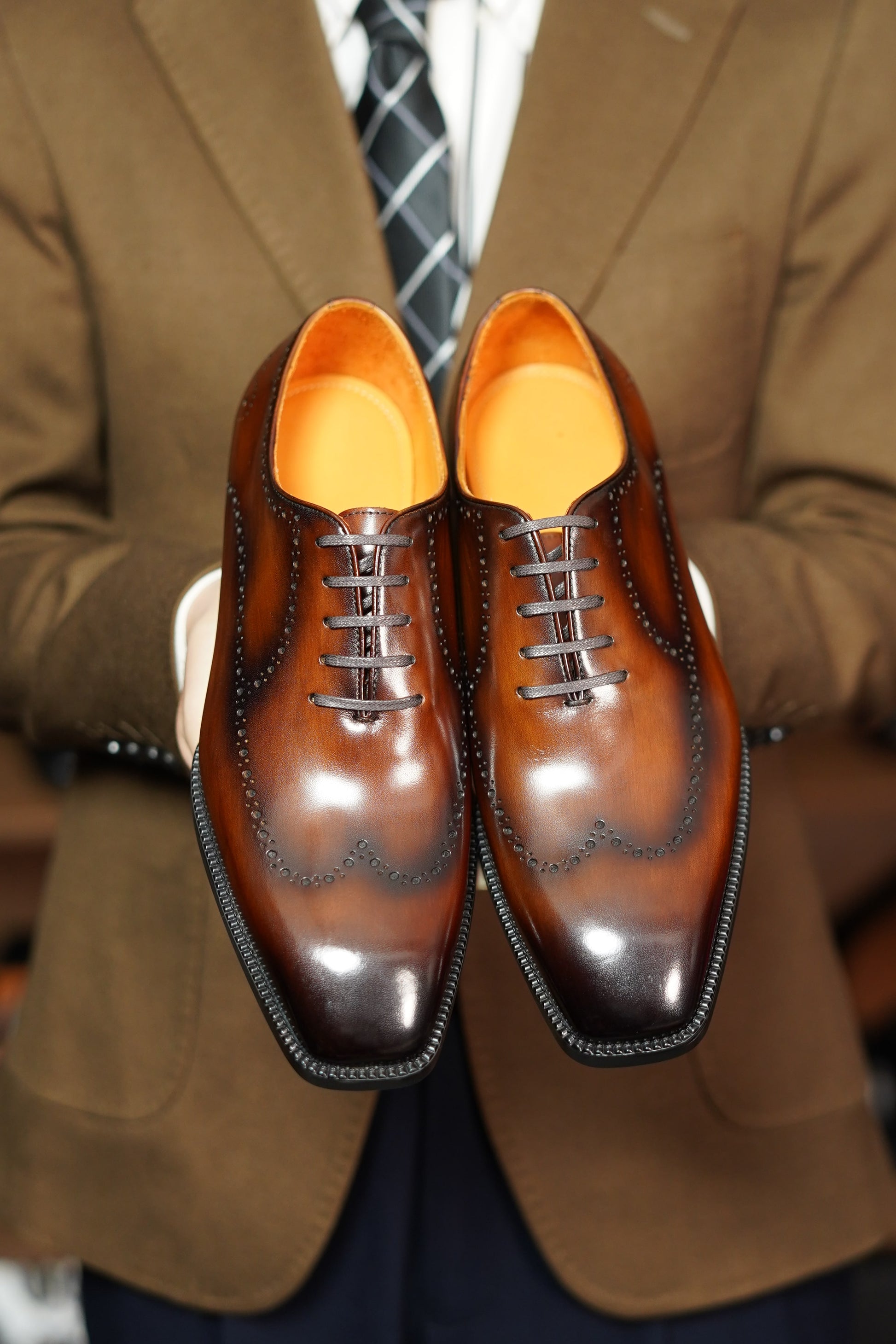Brown dress shoes with orange soles held by a person wearing a brown suit.