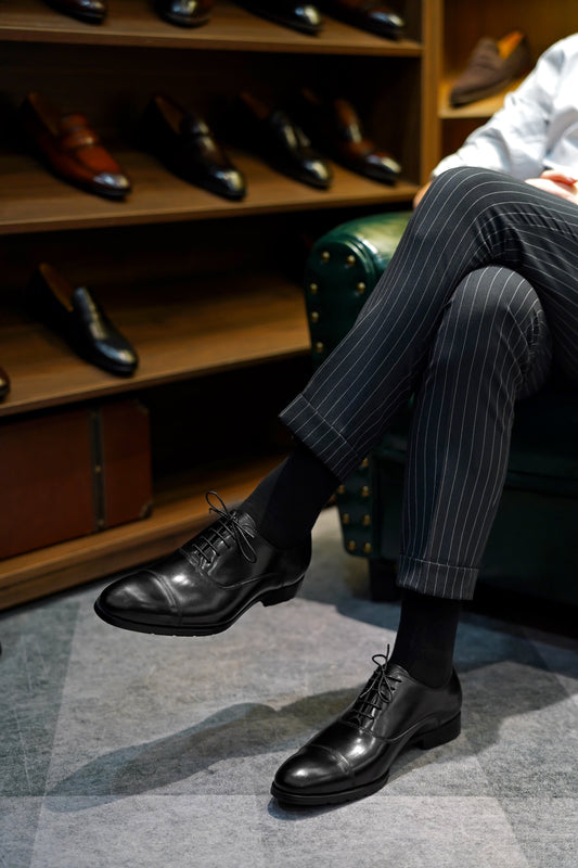 Close-up of men’s black leather cap-toe oxfords, worn with pinstriped trousers, showcasing classic formal design with lightweight and breathable construction for added comfort.