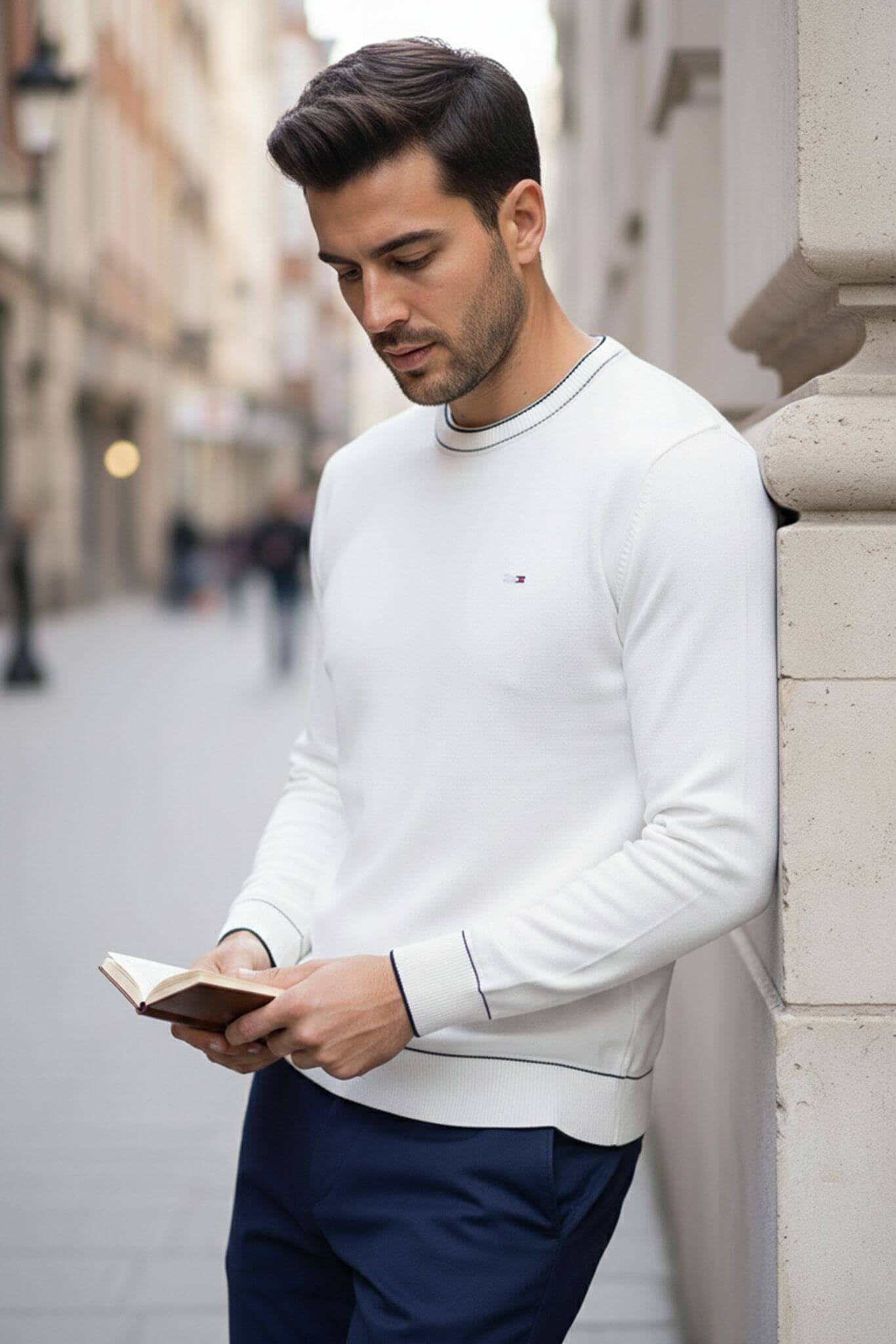 Man in a Slim-fit Crew Neck Sweater with Contrast Piping in White reading a book on a city street.