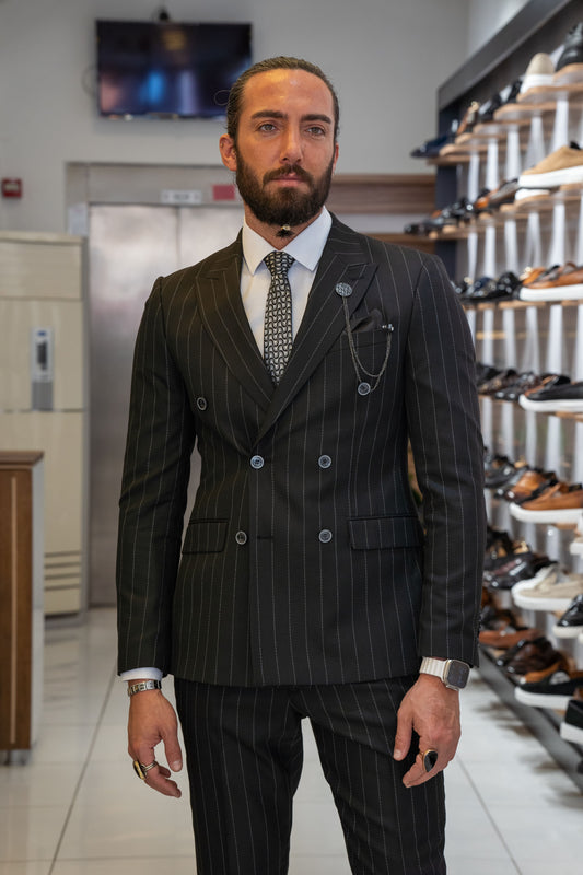 A man standing in a store display window wearing a black double-breasted striped suit with a collar and lapels, paired with black trousers and a dark tie.