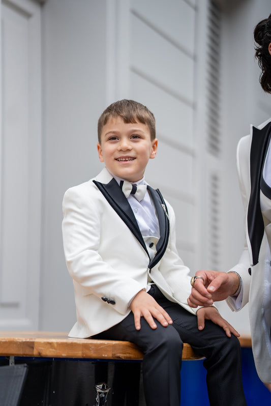 A young boy wearing a white tuxedo suit with black lapels, including a jacket, vest, pants, and a chain pocket, standing next to an adult.