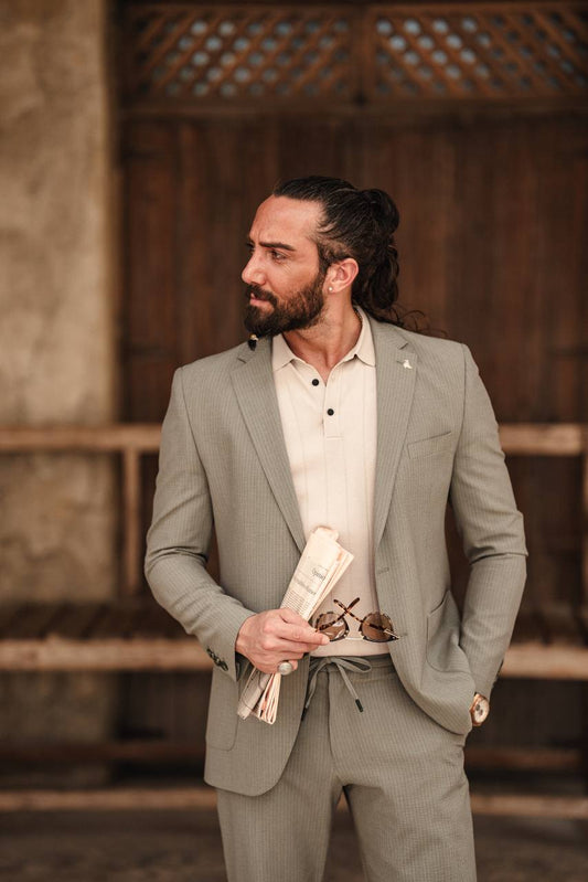 A man wearing a SUITHARBOR Khaki Tailored Suit holding a pair of sunglasses and standing in front of a wooden fence.