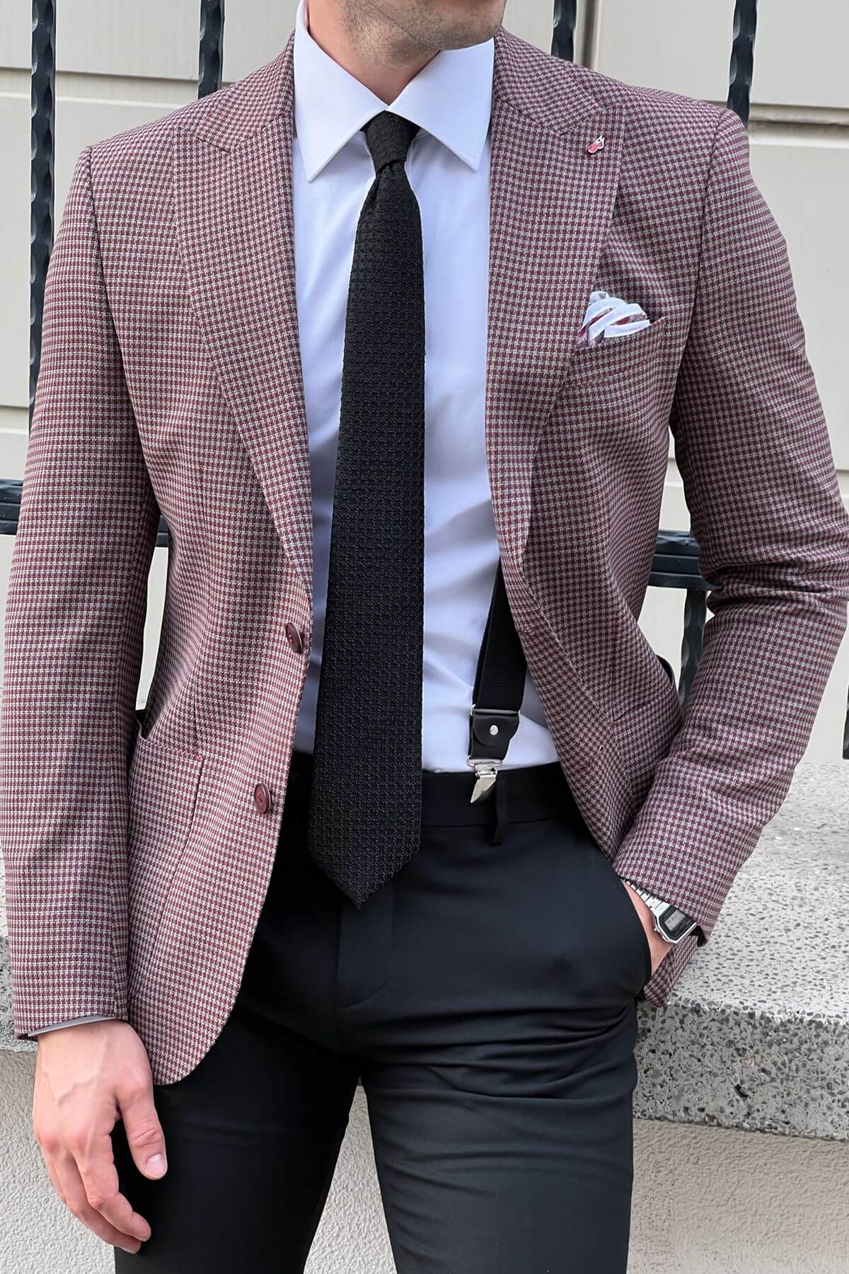 Man wearing a checkered blazer, white shirt, black tie, and suspenders against a neutral background.