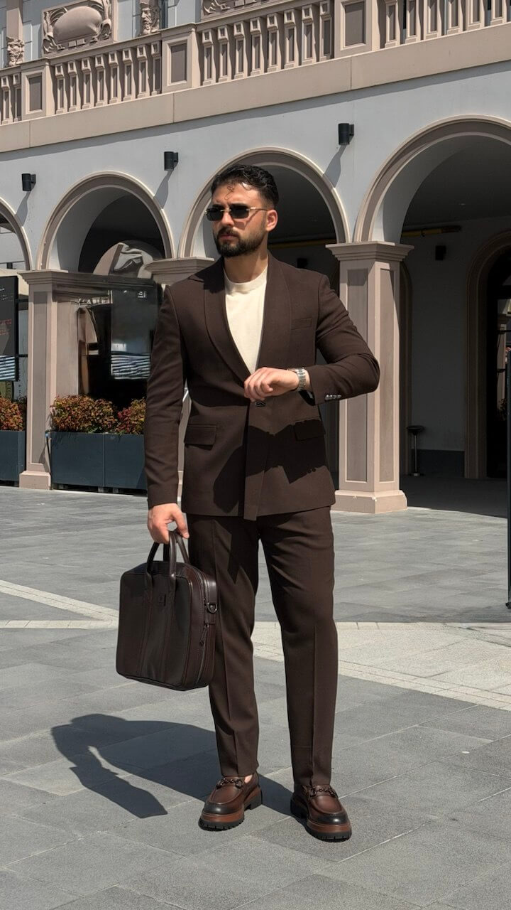 Man in a brown suit holding a briefcase in front of an architectural building.