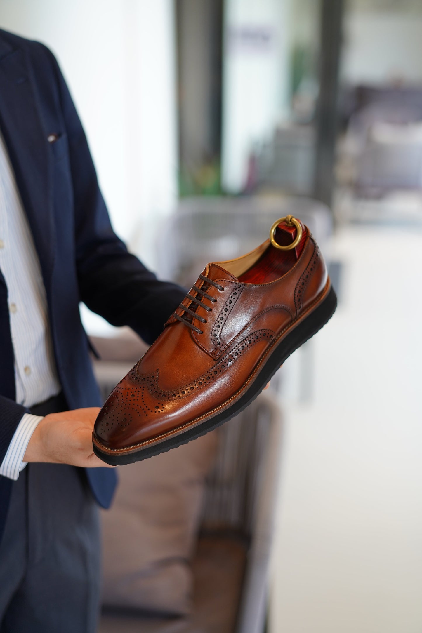 Close-up of a cognac leather brogue shoe with intricate wingtip detailing, held by a man in a navy suit. 