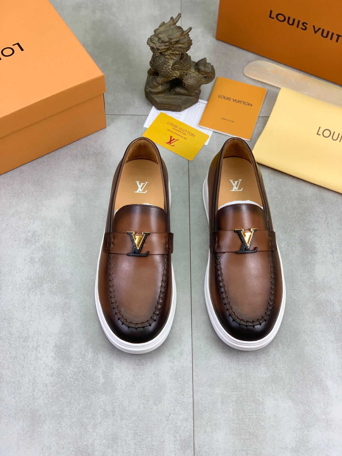A pair of Louis Vuitton brown leather slip-on loafers with white soles, displayed on a gray floor with Louis Vuitton packaging boxes, cards, and a small decorative statue.