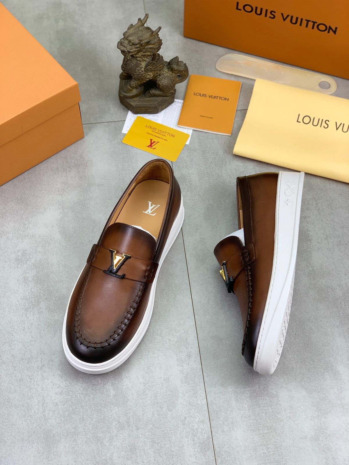 A pair of Louis Vuitton brown leather slip-on loafers with white platform soles, displayed on a gray floor alongside their signature orange and beige Louis Vuitton packaging boxes, dust bag, and cards.
