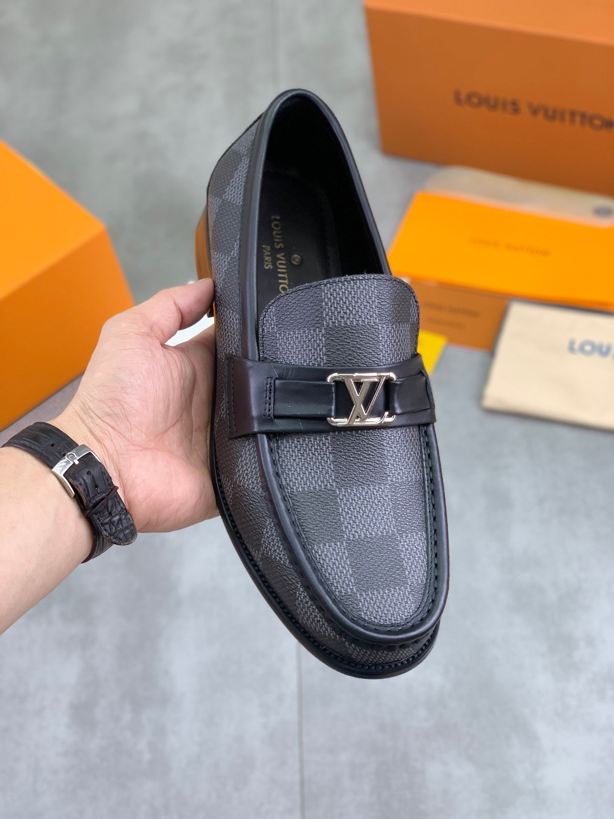 A Louis Vuitton loafers shoe in dark gray and black checkered canvas, held by a hand. The shoe features a signature LV logo on the strap.