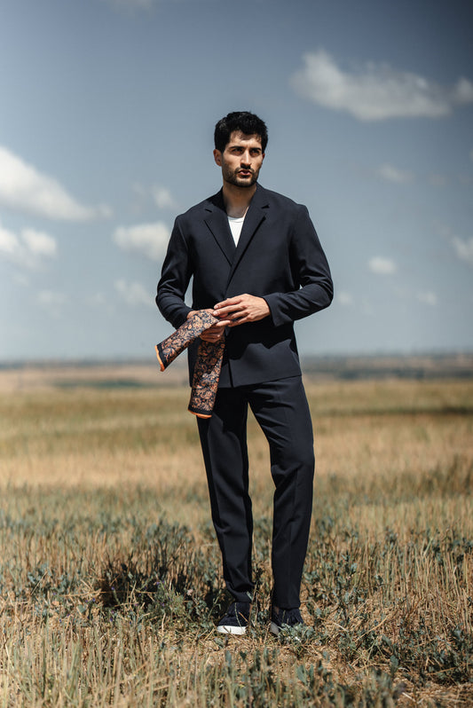 Man in a dark suit holding a patterned pocket square in an open field with a blue sky.