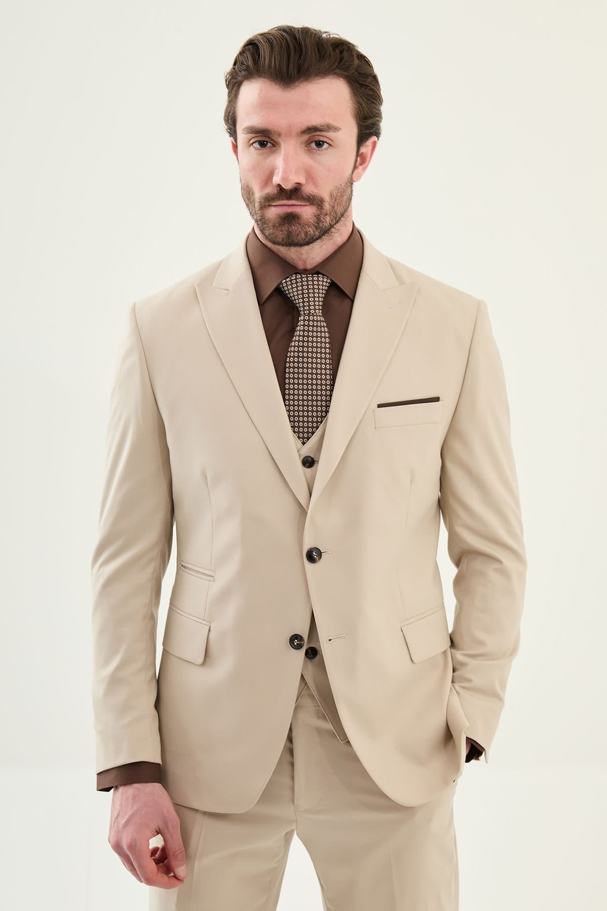 Man wearing a beige suit with a brown shirt and patterned tie on a white background
