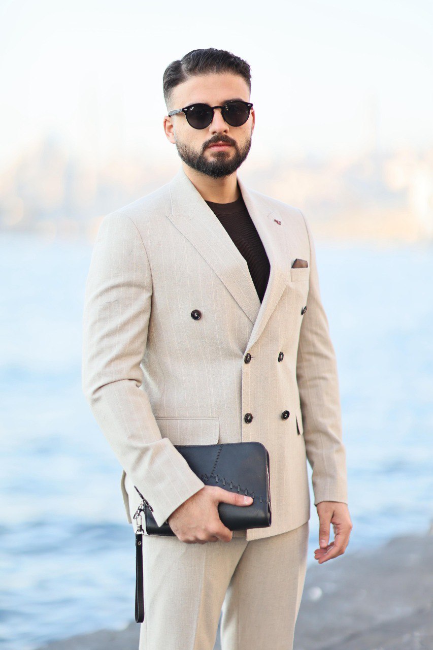 Man in a beige suit with sunglasses standing by a body of water