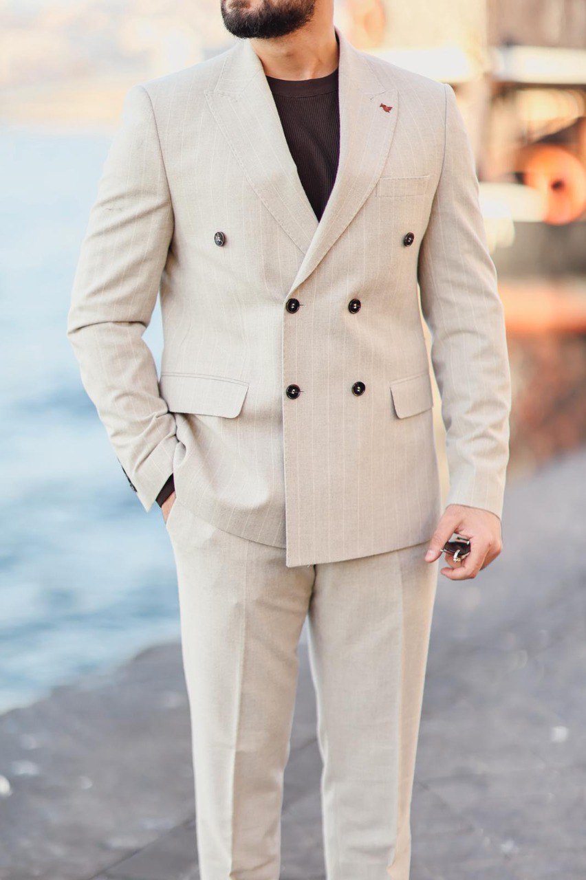 Man wearing a beige suit with black buttons by a waterfront.