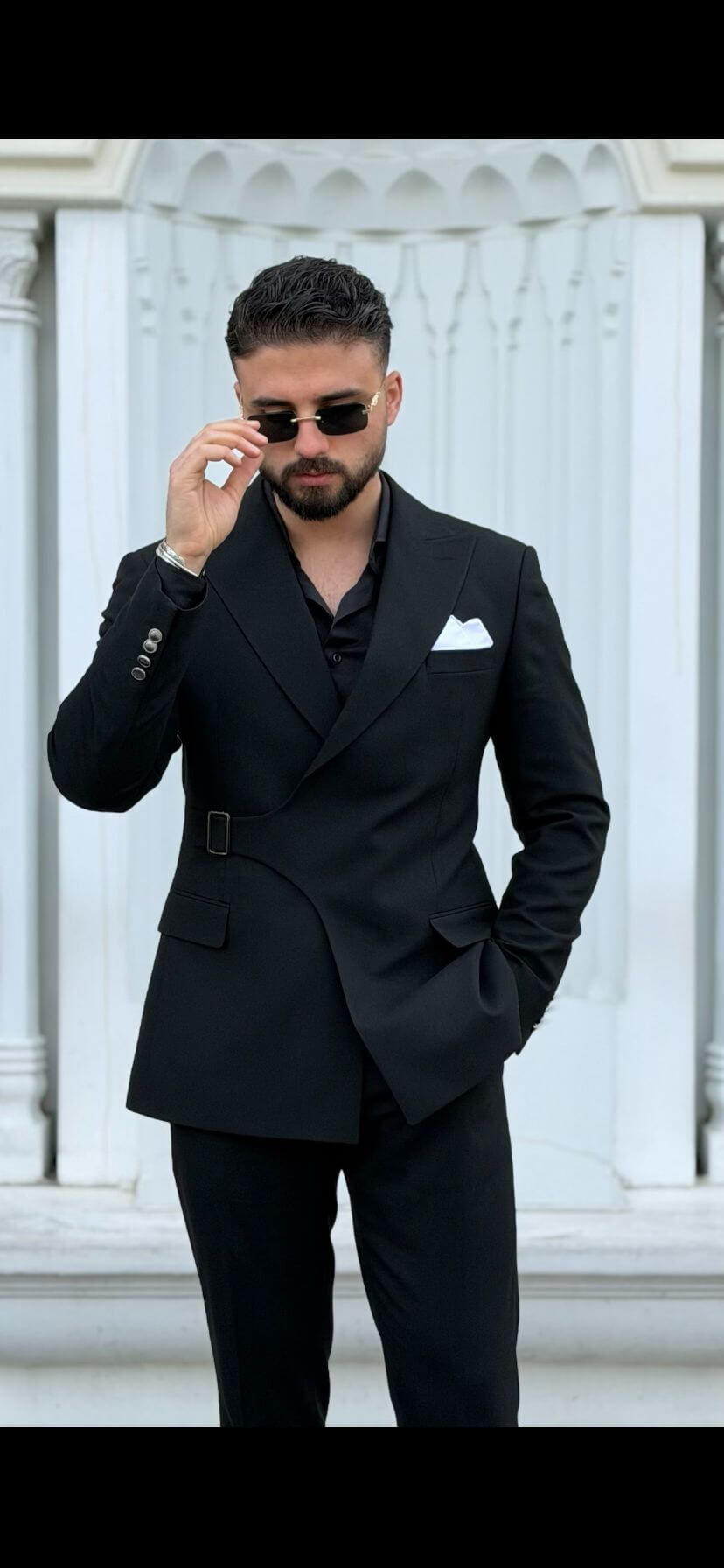 Man wearing a black suit with sunglasses against a white decorative wall.