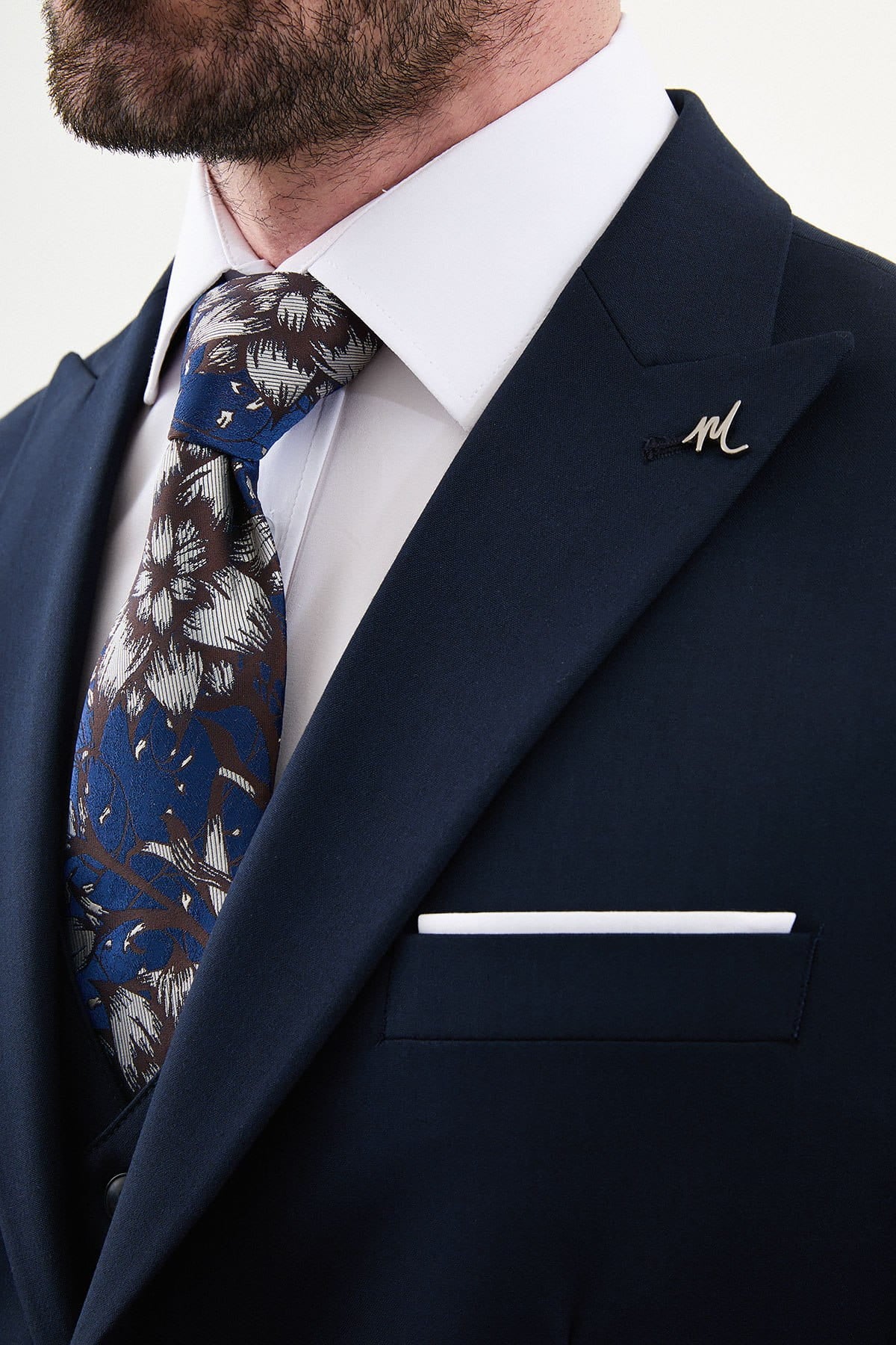 Man wearing a navy suit with a floral tie and a lapel pin on a white background