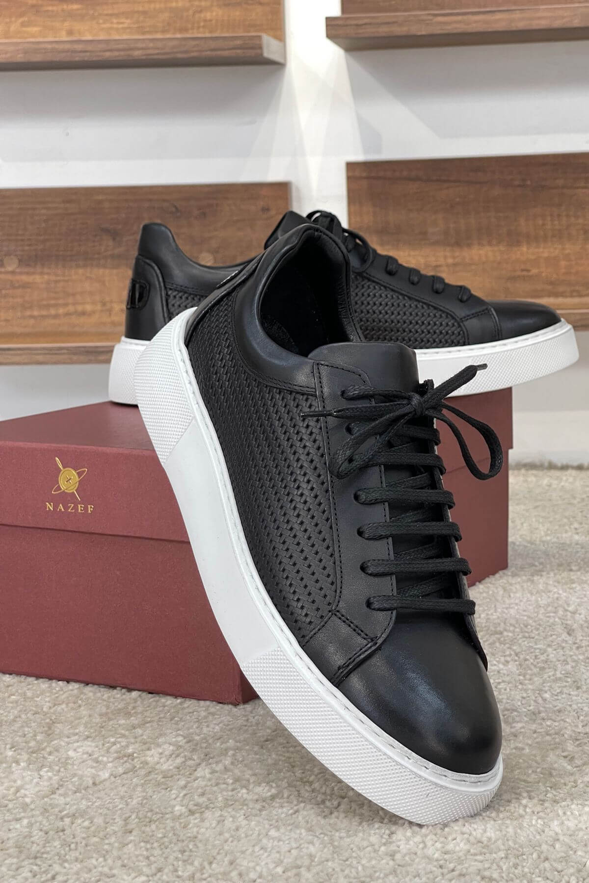 Black sneakers with white soles on a carpeted floor with a wooden shelf in the background.