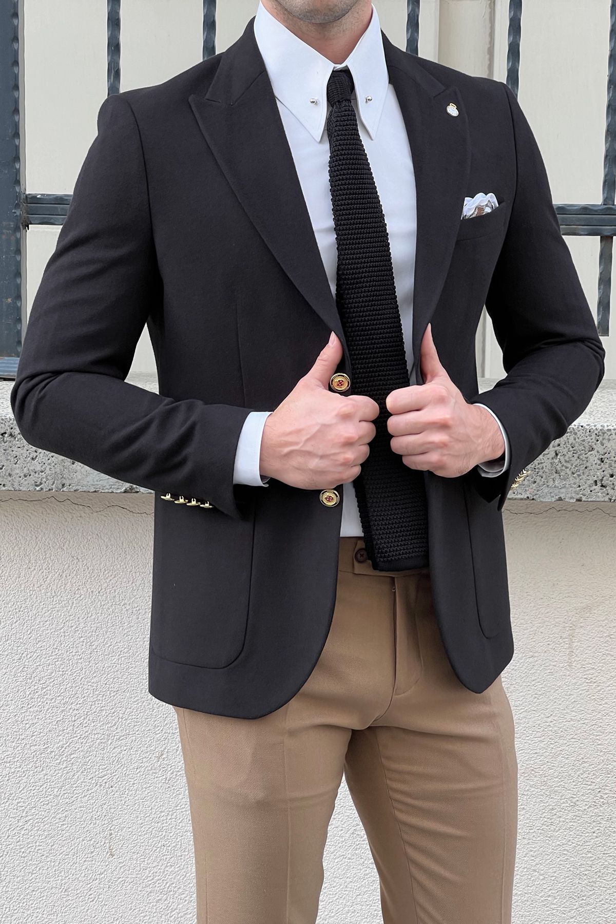 Man wearing a black suit with a white shirt and beige pants, standing against a neutral background.