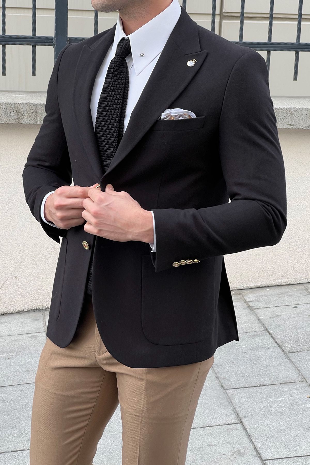 Man wearing a black blazer, white shirt, and beige pants with a patterned tie.