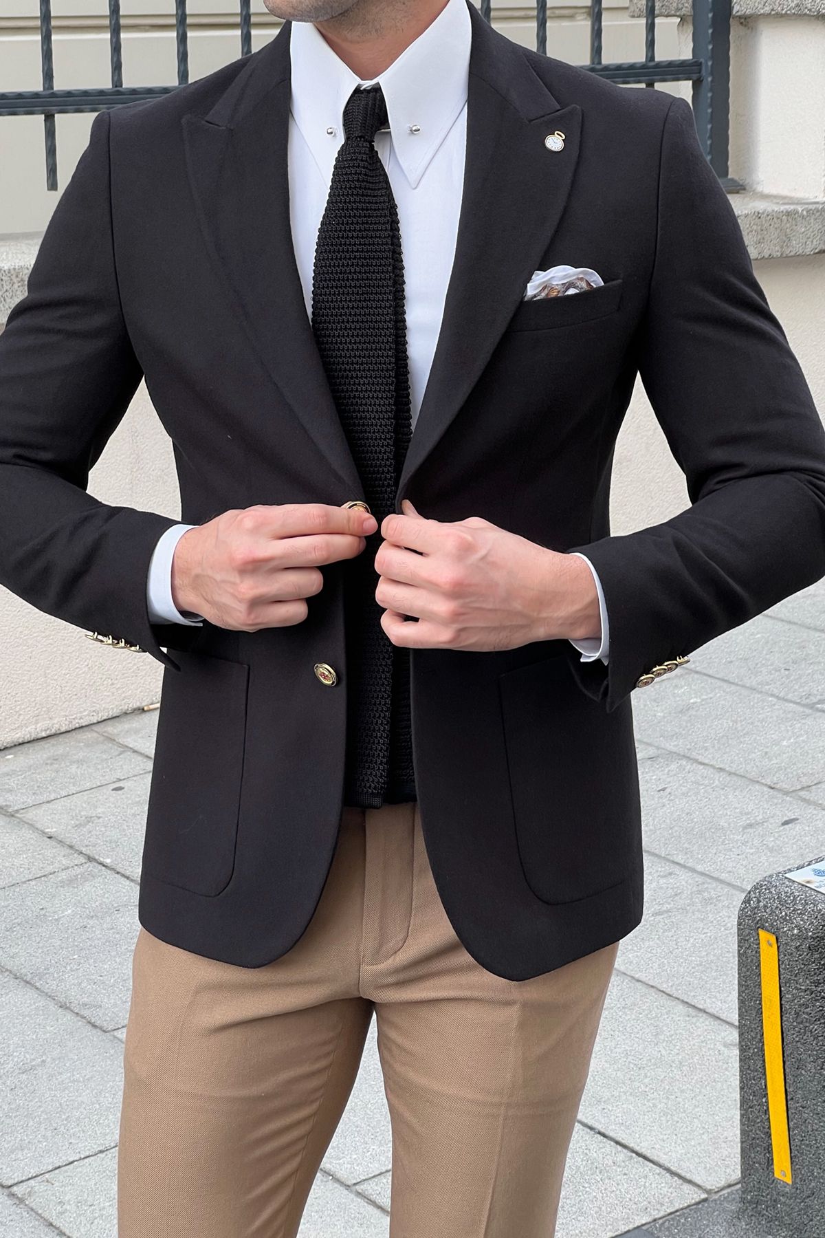Man wearing a black suit jacket, white shirt, and beige pants outdoors.