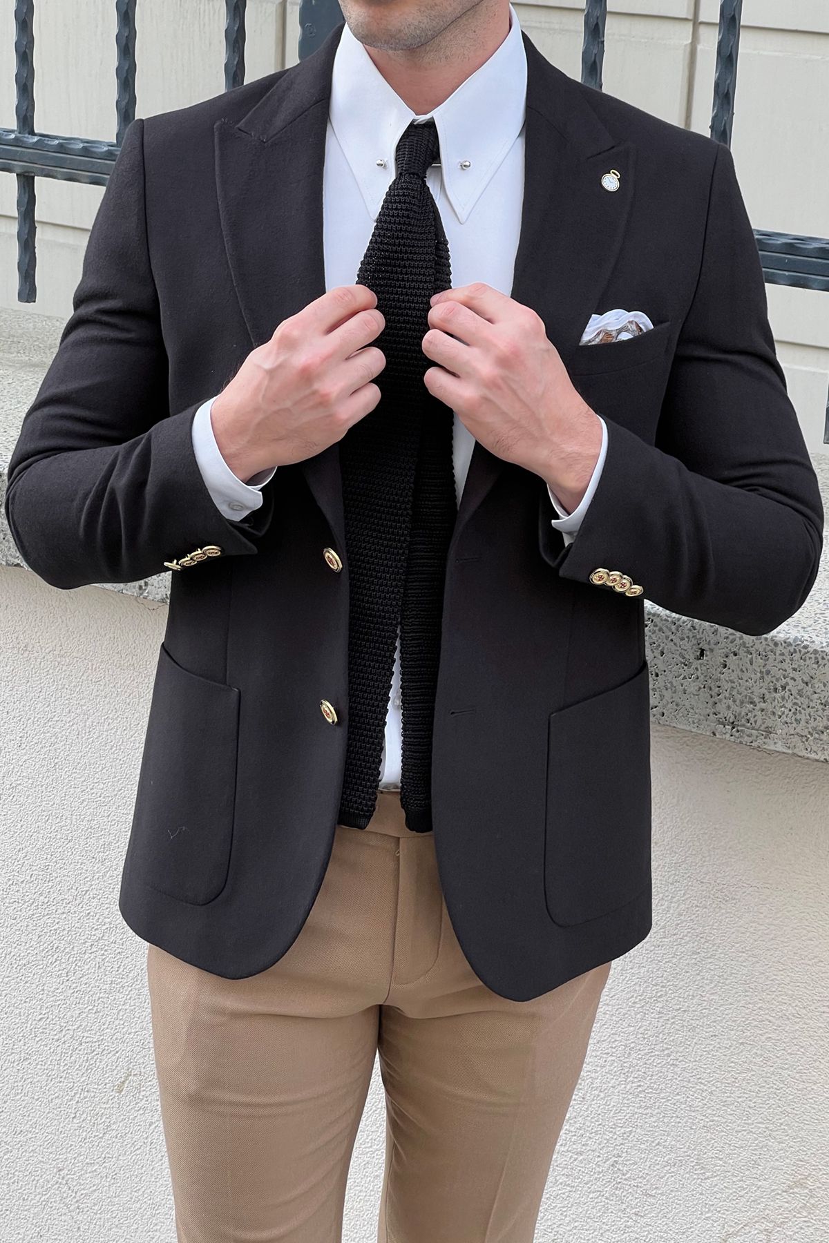 Man adjusting his black tie wearing a black blazer, white shirt, and beige pants.