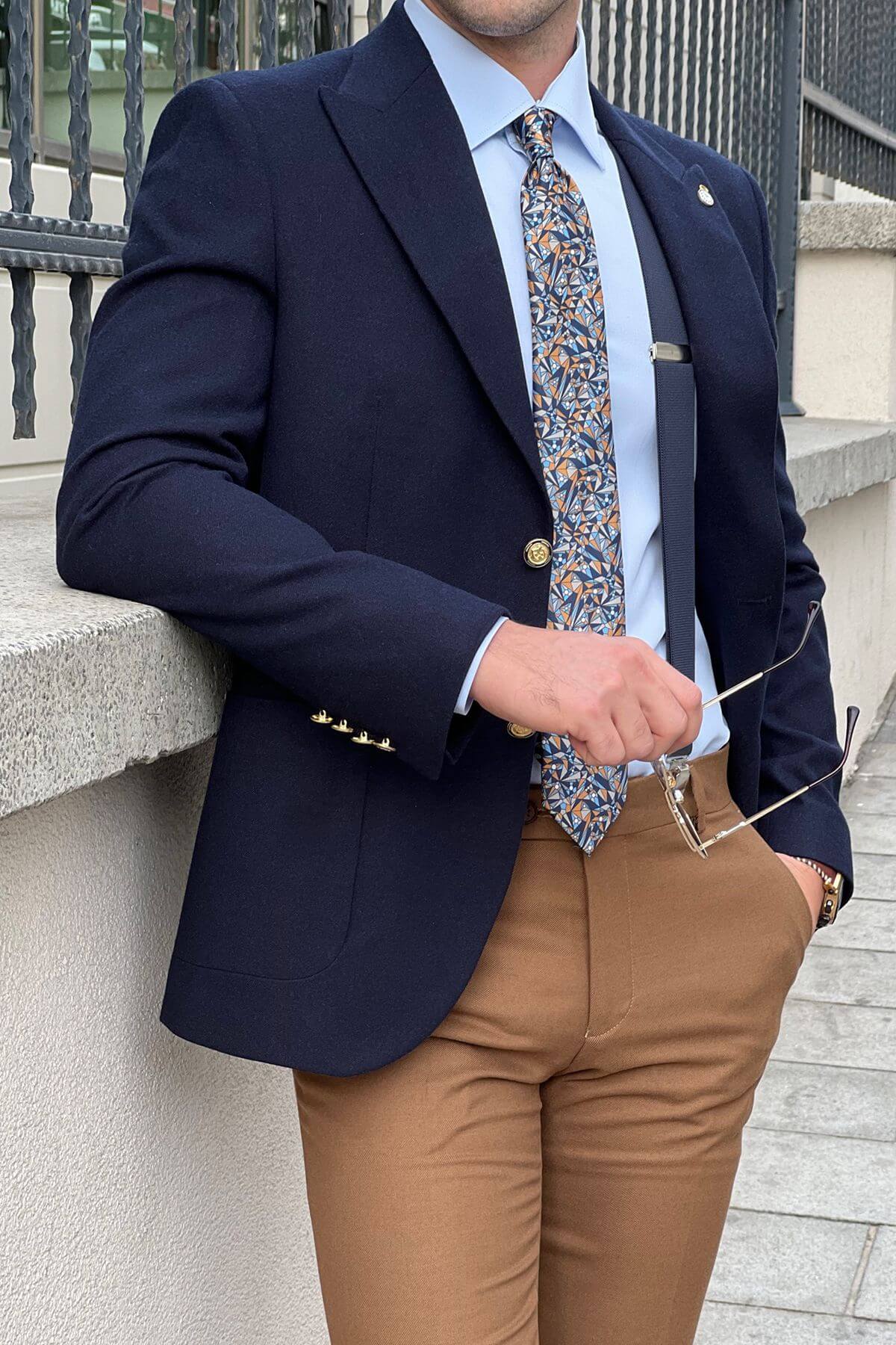 Man wearing a Slim-fit Gold-Buttoned Wool Blend Blazer in Navy Blue, light blue shirt, patterned tie, and brown pants holding glasses.