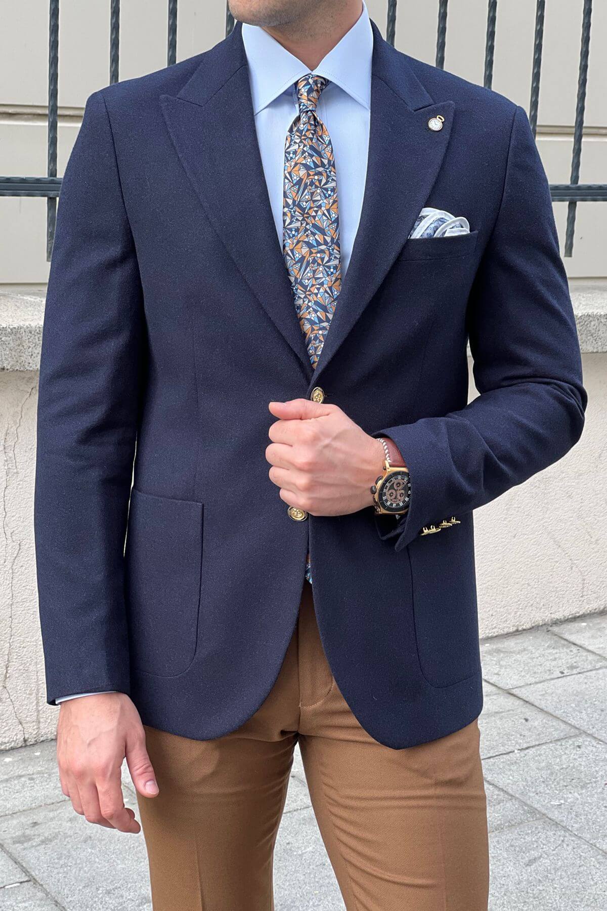 Man wearing a navy blazer, light blue shirt, patterned tie, and brown pants outdoors.