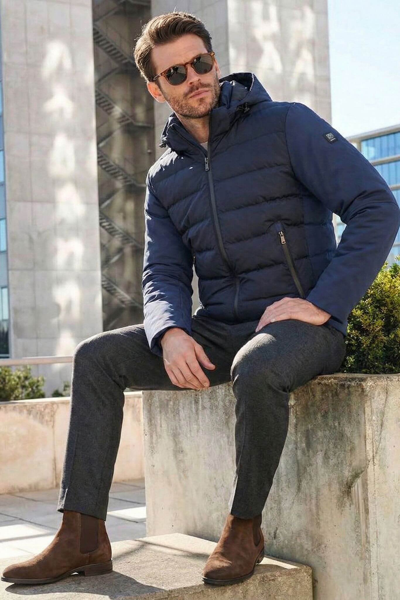 Man wearing a navy puffer jacket sitting on a concrete ledge with a cityscape background.