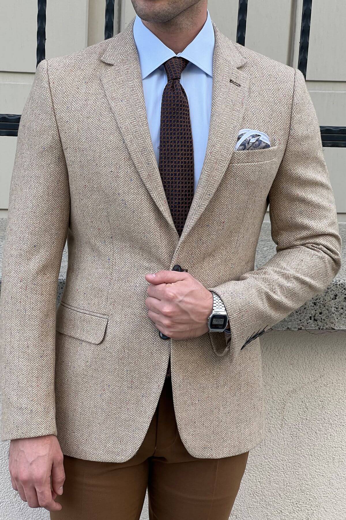 Man wearing a beige suit with a brown tie and pocket square, standing outdoors.