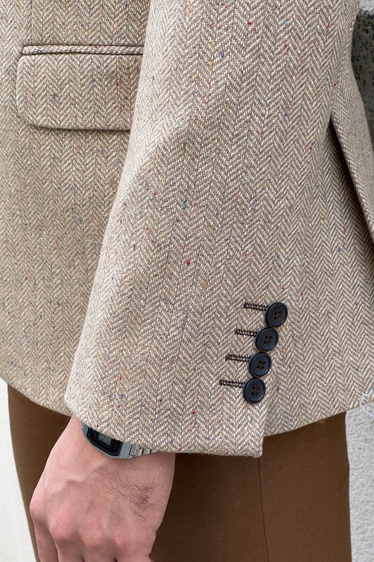 Close-up of a beige herringbone textured coat with buttons on a person wearing brown pants.