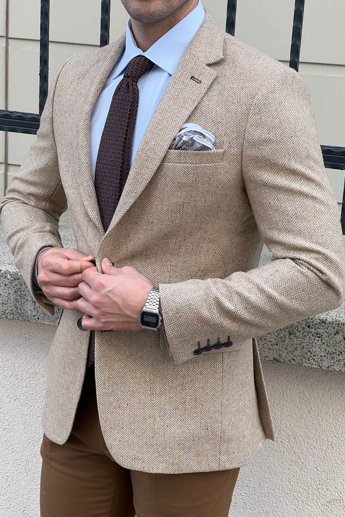 Man wearing a beige blazer with a brown tie and pocket square, standing outdoors.