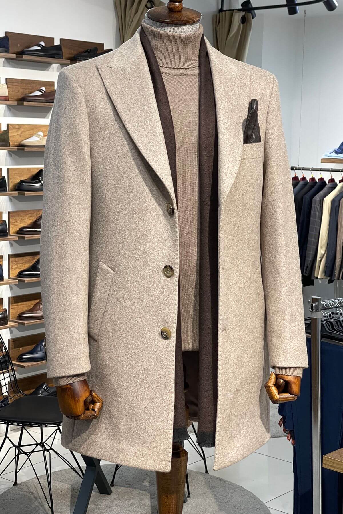 Beige overcoat on a mannequin in a store setting with racks of clothes in the background.