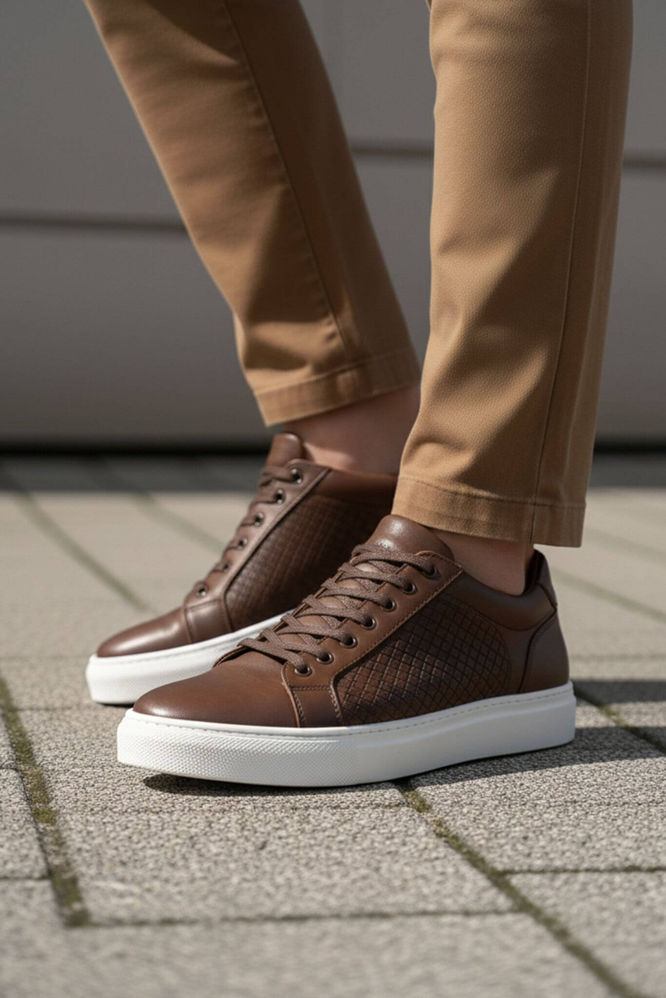 Brown sneakers with white soles worn with beige pants on a pavement.