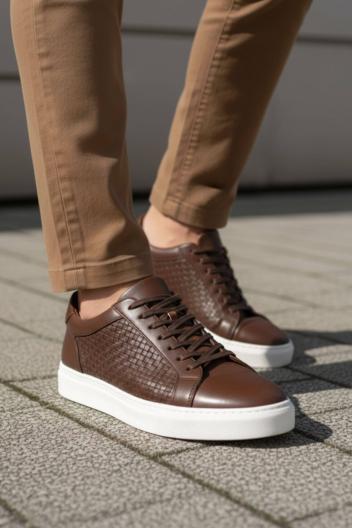Brown leather sneakers with white soles worn with beige pants on a pavement.