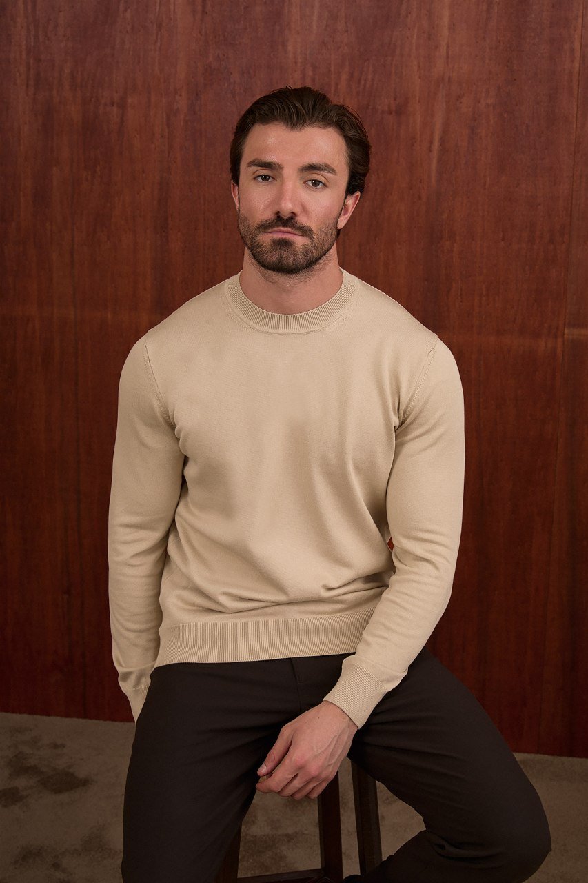 Man seated in a beige crew neck sweater with black trousers