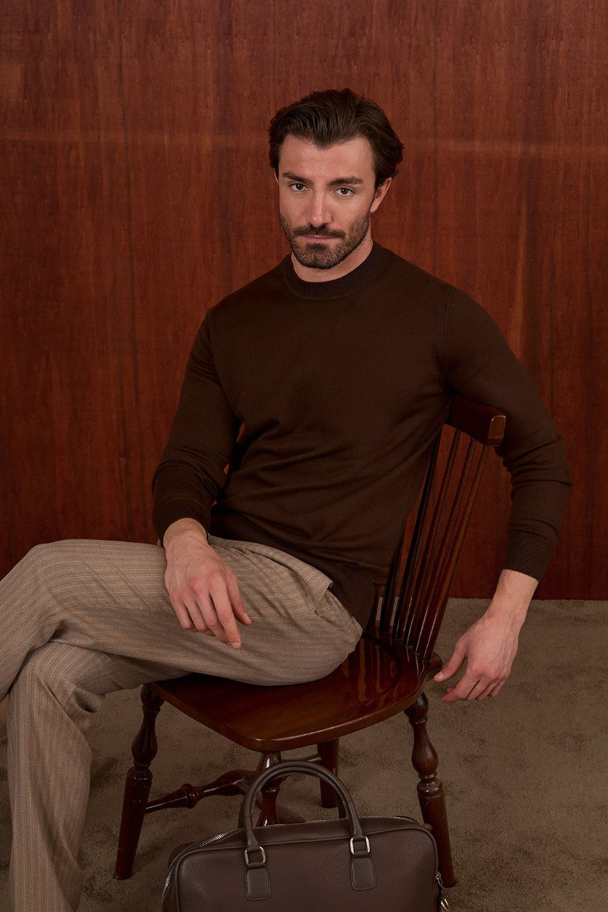 Man seated in a brown crew neck sweater and beige trousers