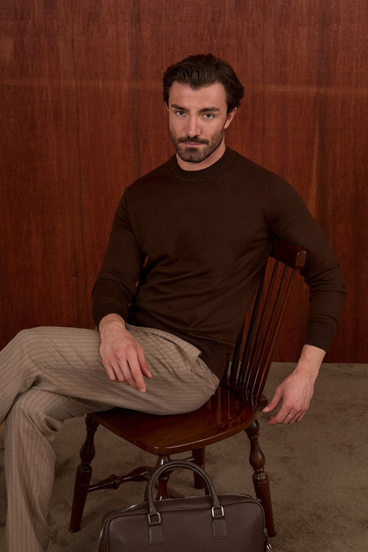Man seated in a brown crew neck sweater and beige trousers