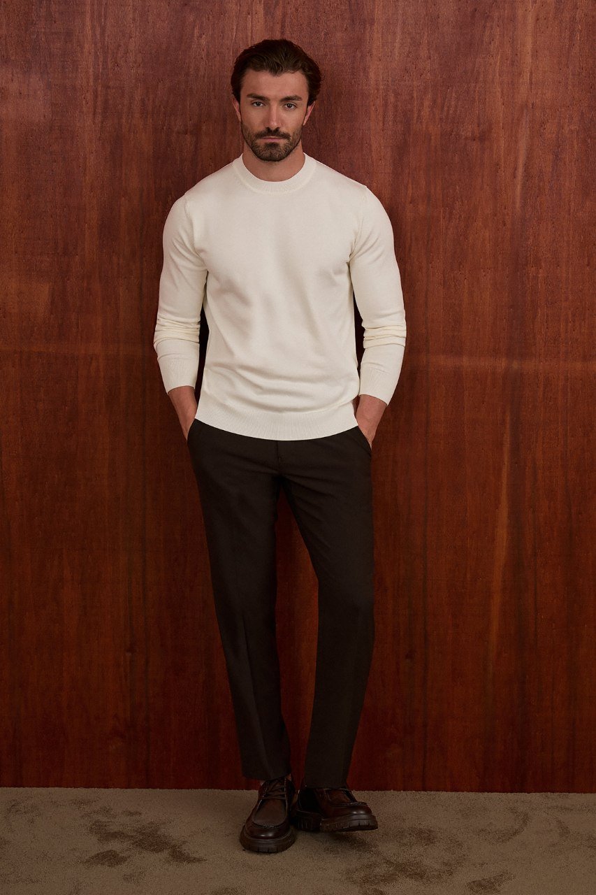 Man wearing an ecru crew neck sweater against a wood-panelled wall