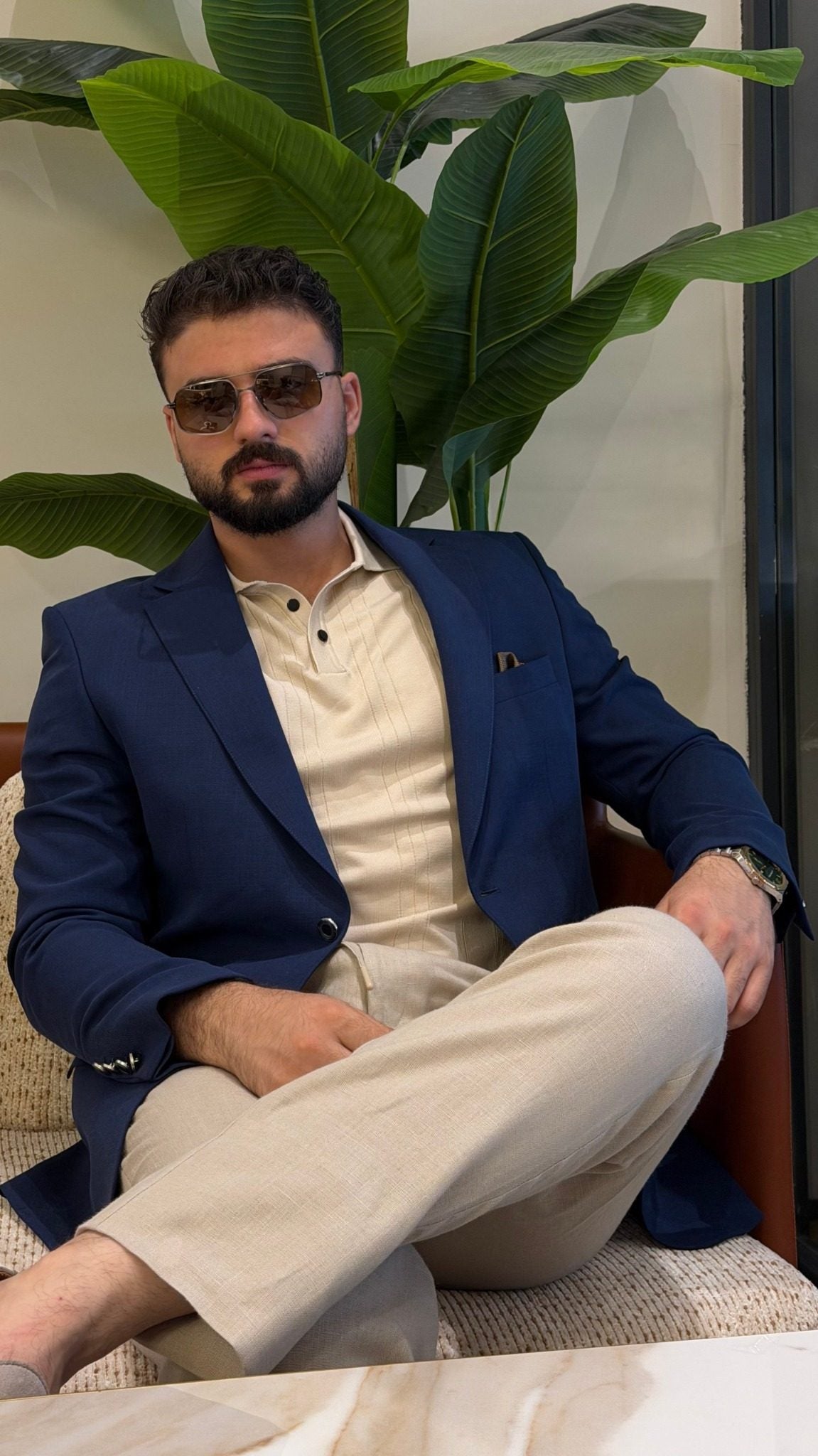 Men's navy blue blazer styled with cream polo and light trousers, seated lifestyle view